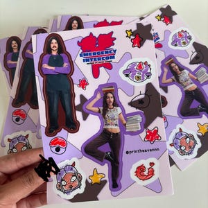 May include: A set of stickers featuring cartoon characters, stars, and a red logo that says "Emergency Intercom" with the text "Headed by Marc Jacobs". The stickers are on a purple background with brown and white accents.