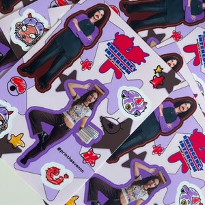 May include: A set of stickers featuring a cartoon character with a purple and pink colour scheme. The stickers are on a purple background with white stars and the text "Emergency Intercom" in a red and white font. The stickers are designed for use on laptops, water bottles, and other surfaces.