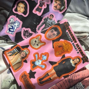 May include: Two sticker sheets with various images, including portraits, cartoon characters, and abstract shapes. The sheets are labeled "Emergency Intercom Sticker Sheet 1" and "Emergency Intercom Sticker Sheet 2". The stickers have a pink and black color scheme.