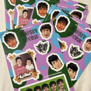 May include: A stack of sticker sheets with a purple background and green and blue circles. The stickers feature a group of young men with dark hair and a photo of two young boys with a cake. The text on the stickers reads "Slushy Noobz" and "Sticker Sheet 2".