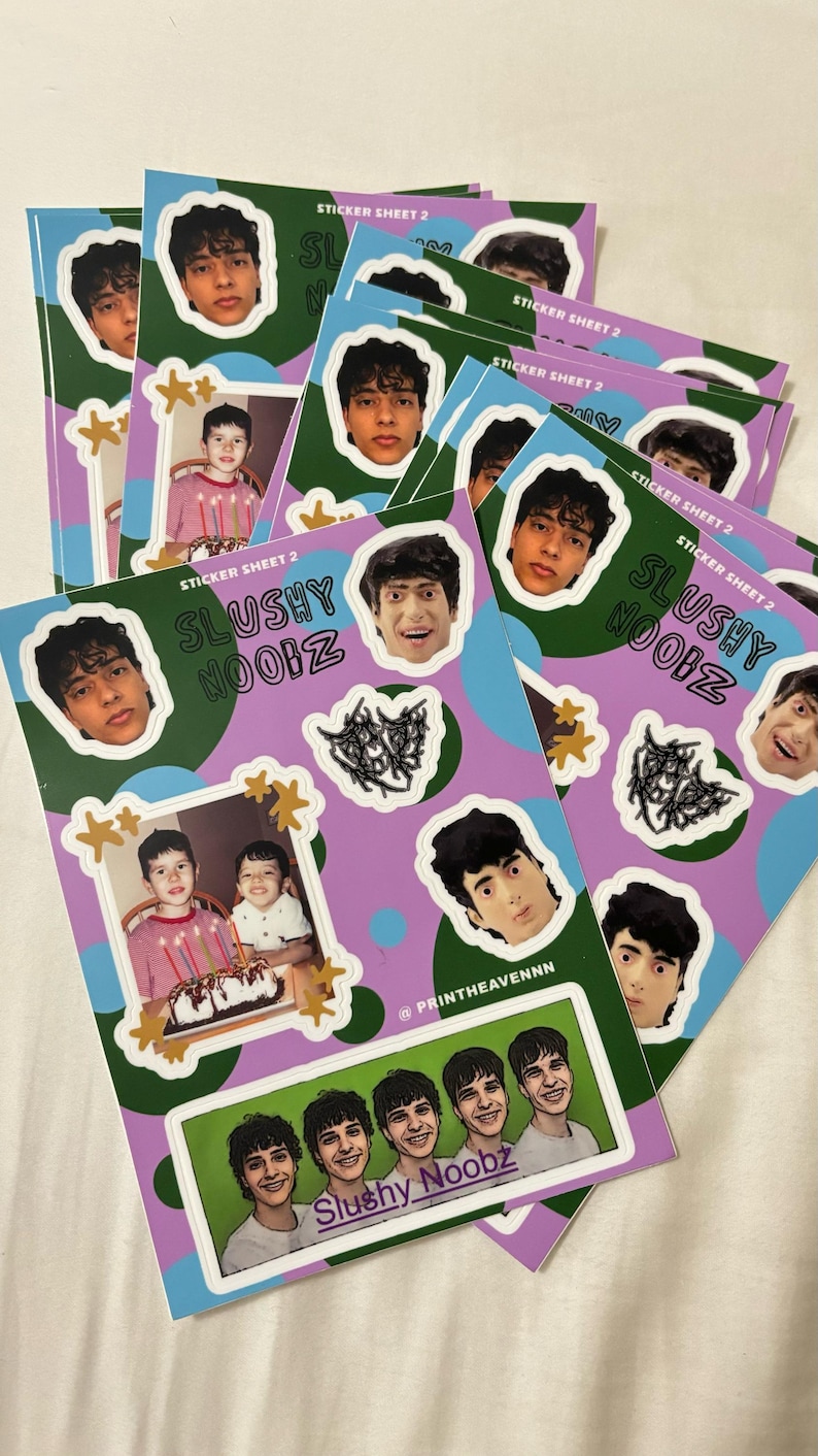 May include: A stack of sticker sheets with a purple background and green and blue circles. The stickers feature a group of young men with dark hair and a photo of two young boys with a cake. The text on the stickers reads "Slushy Noobz" and "Sticker Sheet 2".