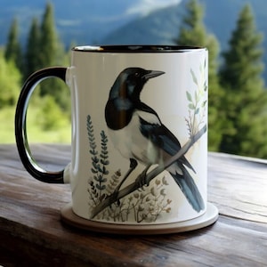 Puede incluir: Una taza de cerámica blanca con un borde y un asa negros presenta una ilustración detallada de una urraca posada en una rama con follaje delicado. El pájaro está representado en tonos negros, blancos y azules.