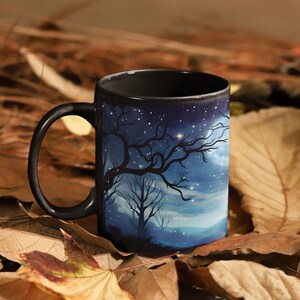 May include: A black ceramic mug with a starry night scene featuring a silhouetted tree and a crescent moon. The mug is sitting on a bed of brown and orange leaves.