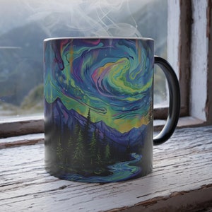 Puede incluir: Una taza negra que cambia de color con un diseño de aurora boreal colorido sobre una cordillera. La taza está llena de una bebida caliente y sale vapor de la parte superior.