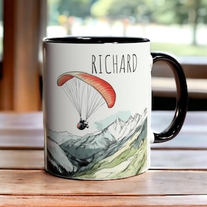 May include: White ceramic mug with a black rim and handle. The mug features a colourful illustration of a paraglider flying over a mountain range. The text "RICHARD" is printed on the mug.