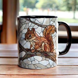 Op de afbeelding: Een keramische koffiemok met een zwart handvat en rand. De mok heeft een glas-in-loodstijl ontwerp van een eekhoorn op een tak, met tinten bruin, oranje en wit.