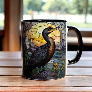 May include: A ceramic mug with a black handle and rim, featuring a colorful stained glass style illustration of a cormorant bird standing on a rock. The background depicts a large yellow sun, blue water, and green foliage in a segmented pattern.