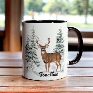 Peut inclure: Mug en céramique blanche avec intérieur et anse noirs. Le mug présente un cerf brun dans une scène de forêt enneigée avec des conifères. Le nom "Jonathan" est imprimé en script noir en dessous.