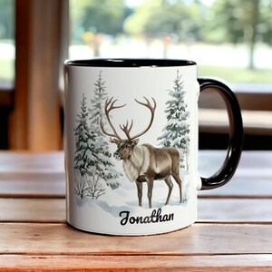 Könnte beinhalten: Weißer Keramikbecher mit schwarzem Innenraum und Henkel. Der Becher zeigt eine Winterszene mit einem Rentier und immergrünen Bäumen. Der Name "Jonathan" ist in schwarzer Schrift am Boden des Bechers aufgedruckt.