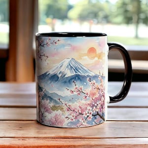 May include: A ceramic mug with a black handle and interior. The mug features a watercolor painting of Mount Fuji with cherry blossoms and a setting sun. The background is a soft blend of pink, blue, and yellow hues.