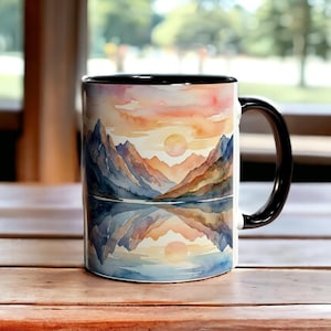 May include: A ceramic coffee mug with a black handle and interior. The mug features a watercolor landscape design of mountains, a lake, and a sunset in shades of orange, yellow, and blue. The mug is sitting on a wooden surface.