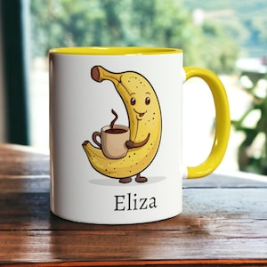 Op de afbeelding: Een witte keramische mok met een gele rand en handvat. De mok heeft een cartoonbanaan die een kopje koffie vasthoudt en de naam "Eliza" staat eronder gedrukt.
