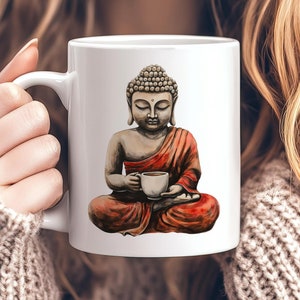 May include: White ceramic mug with a colorful illustration of Buddha sitting in a meditative pose, holding a small cup. The Buddha is wearing a red robe and has a serene expression.
