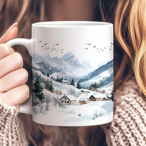 May include: White ceramic mug featuring a winter landscape scene with snow-covered mountains, trees, and houses. The design includes a flock of birds in the sky. The mug has a curved handle and is held by a person wearing a brown sweater.