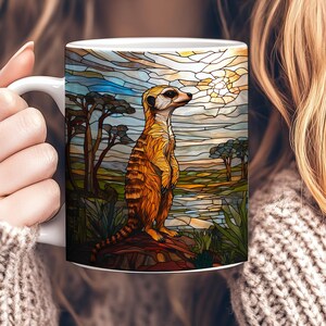 Peut inclure: Une tasse en céramique blanche avec un motif de vitrail représentant un suricate debout sur un rocher devant un lac. Le suricate est brun et jaune avec une queue noire. L'arrière-plan est un ciel bleu avec un soleil jaune.