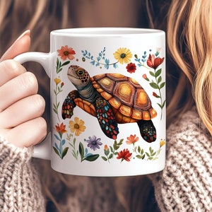 Könnte beinhalten: Weiße Keramiktasse mit einer farbenfrohen Schildkrötenillustration und einem floralen Rand. Die Schildkröte hat einen orangefarbenen und braunen Panzer. Die Blumen umfassen rote, gelbe und violette Blüten. Die Tasse hat einen gebogenen Griff.