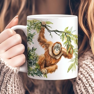 Peut inclure: Une tasse en céramique blanche avec une illustration aquarelle d'un orang-outan suspendu à une branche d'arbre. L'orang-outan est brun et a un fond vert feuillu.