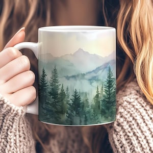 May include: White ceramic mug featuring a watercolor landscape of a mountain range and evergreen trees in shades of green and blue. The mug has a white handle and is held by a person wearing a beige sweater.