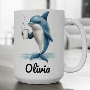 May include: White ceramic mug with a blue dolphin illustration holding a cup of coffee. The dolphin is surrounded by blue bubbles and the mug is personalized with the name "Olivia".