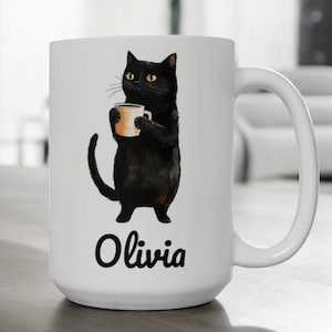 Könnte beinhalten: Eine weiße Keramiktasse mit einer Illustration einer schwarzen Katze, die eine Kaffeetasse hält. Die Katze schaut nach rechts und der Text "Olivia" ist unter der Katze aufgedruckt.