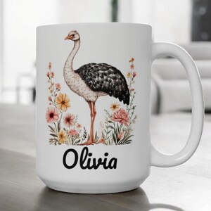 Puede incluir: Una taza de cerámica blanca con una ilustración impresa de un avestruz rodeado de flores coloridas. El nombre Olivia está impreso en letras cursivas negras debajo del avestruz. La taza tiene un asa curva en el lado derecho.