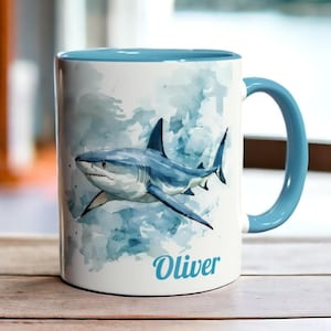 May include: A white ceramic mug with a blue rim and handle. The mug features a watercolour illustration of a shark swimming in the ocean. The name "Oliver" is written in blue cursive at the bottom of the mug.