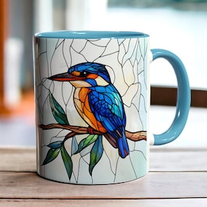 Könnte beinhalten: Keramikbecher mit blauem Griff und Innenraum. Die Außenseite zeigt eine Buntglas-Illustration eines Eisvogels, der auf einem Ast mit grünen Blättern sitzt. Der Vogel hat blaue, orange und weiße Federn.