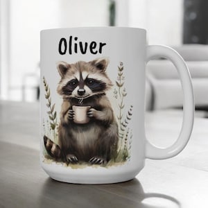 May include: White ceramic mug with a black handle. The mug features a cartoon raccoon holding a cup of coffee and surrounded by green foliage. The text "Oliver" is printed in black on the mug.