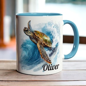 Könnte beinhalten: Eine weiße Keramiktasse mit blauem Rand und Henkel. Die Tasse zeigt eine Aquarell-Illustration einer Meeresschildkröte, die in einer Welle schwimmt. Der Text "Oliver" ist auf die Tasse gedruckt.