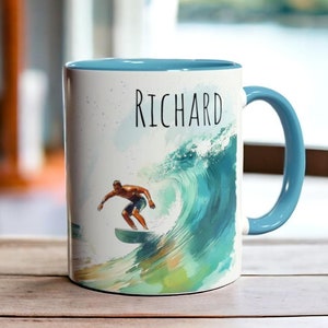 Op de afbeelding: Witte keramische mok met een blauw interieur en handvat. De mok heeft een aquarel illustratie van een surfer op een golf, met de naam "Richard" in zwart schrift. Het ontwerp bevat tinten blauw, groen en wit.