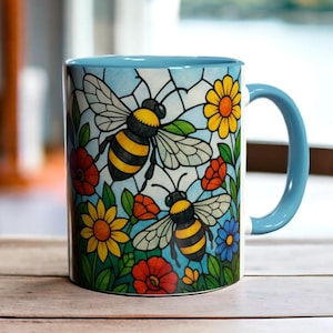 May include: A blue ceramic mug with a handle features a colorful stained glass design of two bees flying among flowers. The bees are yellow and black, and the flowers are red, yellow, blue, and green.