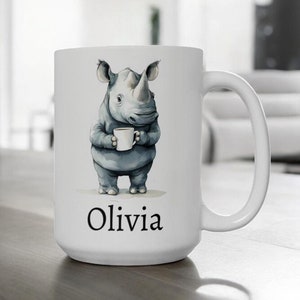 Op de afbeelding: Witte keramische mok met een cartoon neushoorn die een kopje koffie vasthoudt. De mok heeft de naam "Olivia" onderaan gedrukt.