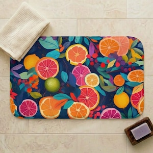 Peut inclure: Un tapis de bain rectangulaire avec un motif d'agrumes vibrant. Le tapis présente des oranges, pamplemousses et citrons tranchés dans des tons orange, rose et jaune, sur un fond bleu foncé avec des feuilles vertes. Une serviette blanche et un porte-savon en bois sont également présents.