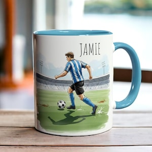 Op de afbeelding: Witte keramische mok met een blauwe binnenkant en handvat. De mok heeft een illustratie van een voetballer in een blauw-wit gestreept tenue, die een bal op een groen veld trapt. De naam "JAMIE" staat boven de speler.