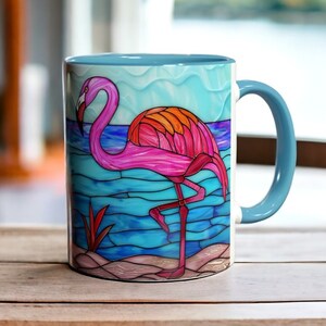 May include: A ceramic mug with a flamingo design. The flamingo is pink, orange, and red, set against a blue water background. The mug has a light blue handle and interior. The mug is on a wooden surface.