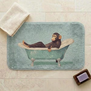 May include: A rectangular light blue bath mat featuring a printed illustration of a brown chimpanzee relaxing in a clawfoot bathtub. The mat has a soft, plush texture and is displayed alongside a folded cream-colored towel and a purple soap bar.