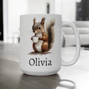Puede incluir: Taza de café de cerámica blanca con una ilustración de dibujos animados de una ardilla sosteniendo una taza de café. La taza tiene el nombre "Olivia" impreso en la parte inferior.