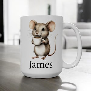 Puede incluir: Taza de café de cerámica blanca con una ilustración de dibujos animados de un ratón marrón sosteniendo una taza de café. La taza tiene el nombre "James" impreso en la parte inferior.