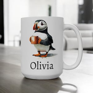 Op de afbeelding: Witte keramische mok met een cartoonillustratie van een papegaaiduiker die een kopje koffie vasthoudt. De mok heeft de naam "Olivia" onderaan gedrukt.