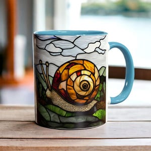 Könnte beinhalten: Eine weiße Keramiktasse mit blauem Rand und Henkel. Die Tasse zeigt ein Buntglasdesign mit einer Schnecke mit einer gelben, orangefarbenen und braunen Schale, die auf grünen Blättern kriecht.