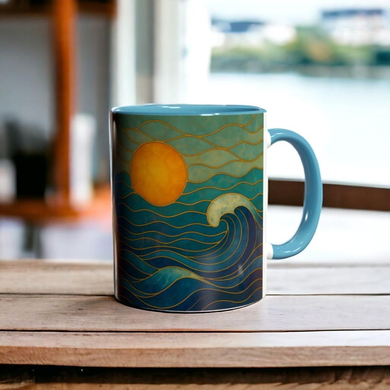 May include: A blue and white ceramic mug with a colorful illustration of a sun setting over a wavy ocean. The mug has a blue handle.
