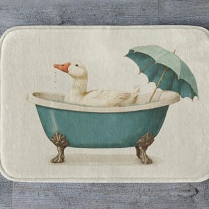May include: A rectangular bath mat with a charming illustration of a white duck in a teal claw-foot bathtub, under a teal umbrella. The mat has a cream background and is suitable for a bathroom. The bathtub is reminiscent of a vintage style.