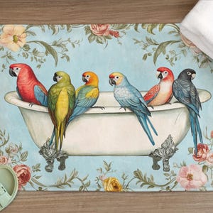 Op de afbeelding: Badmat met een vintage illustratie van zes kleurrijke papegaaien op een klauwvoet badkuip. De mat heeft een lichtblauwe achtergrond met bloemen- en wijnstokaccenten. Een witte handdoek is zichtbaar.