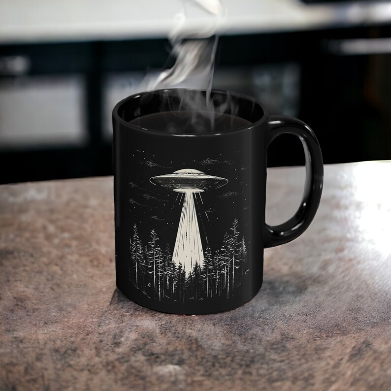 May include: Black coffee mug featuring a white illustration of a UFO hovering over a forest at night. The mug has a black handle.