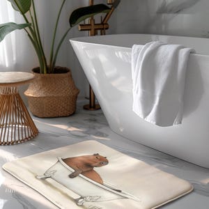 May include: A rectangular bath mat featuring a whimsical illustration of a capybara relaxing in a bathtub. The mat has a beige background and is placed on a marble floor. A white bathtub and towel are in the background.