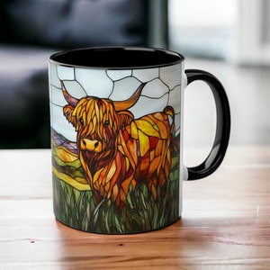 May include: A black coffee mug with a white design of a Highland cow in a stained glass style. The cow is brown and orange with a white face and horns. The background is a green field with a blue sky.