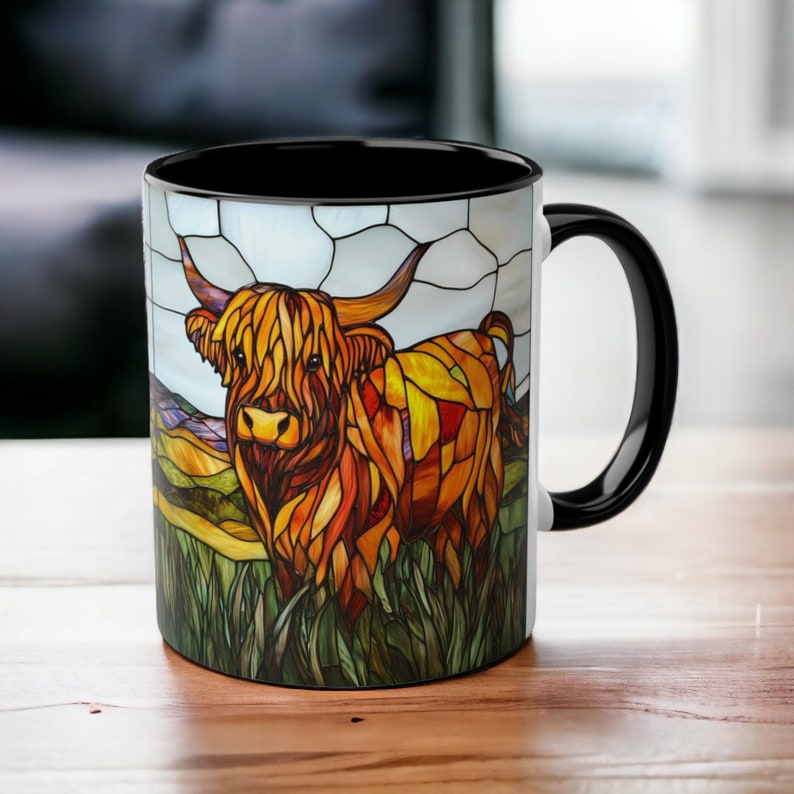 May include: A black coffee mug with a white design of a Highland cow in a stained glass style. The cow is brown and orange with a white face and horns. The background is a green field with a blue sky.