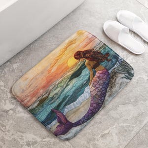 Può includere: Un tappetino da bagno con un'illustrazione ad acquerello di una sirena con squame viola e blu seduta su una spiaggia con un tramonto sullo sfondo.