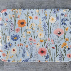 Könnte beinhalten: Rechteckige Badematte mit floralem Design. Die Matte hat einen hellblauen Hintergrund mit einem Muster aus bunten Wildblumen in Orange-, Rosa-, Gelb- und Blautönen. Die Matte hat abgerundete Ecken.