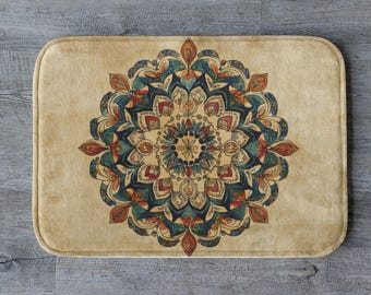 May include: Rectangular beige bath mat featuring a detailed mandala design in shades of teal, red, and orange. The floral pattern is centered on the mat, creating a symmetrical and decorative element. The mat has rounded corners.